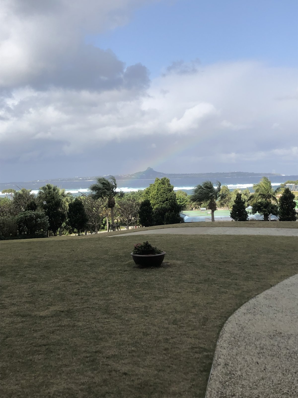 沖縄本島・本部町のホテルオリオンモトブリゾート＆スパから望む絶景。ヤシの木と緑の庭・伊江島の城山・海上にかかる虹が映える夏沖縄のリゾート風景