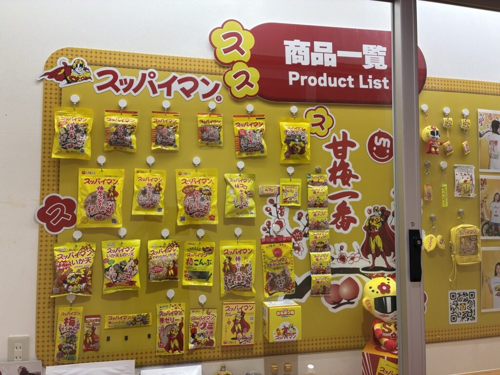 上間菓子店本店の商品一覧ボード。スッパイマン甘梅一番をはじめとする黄色いパッケージの商品が並ぶ