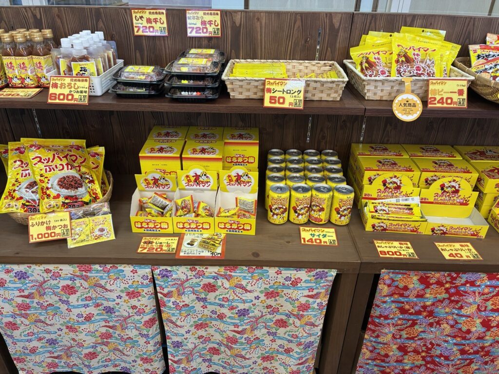 スッパイマン本店の売店の商品棚。ふりかけ・サイダー・柿ピー一番などスッパイマン味の商品が値札つきで並ぶ